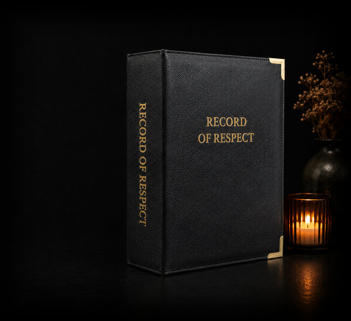 Record of Respect black leatherette memory album with gold corners and spine lettering, standing beside a lit candle and a vase of dried flowers